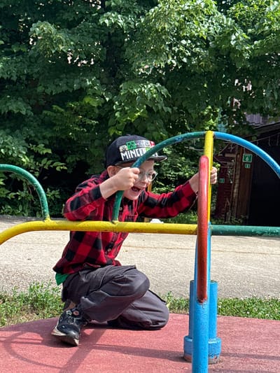 A young boy wearing a red and black checkered shirt, dark pants, and a black baseball cap is on a playground in Yunost', Russia. He is kneeling and holding onto a colorful metal play structure that has yellow, blue, green, and red bars. The boy is laughing with his mouth open and is looking towards the viewer. The playground appears to be outdoors with trees and foliage in the background, suggesting it is a sunny day. The ground beneath him is red and textured, typical of a playground surface. There are also visible hints of concrete and grass. A wooden building with windows is visible behind the play structure.