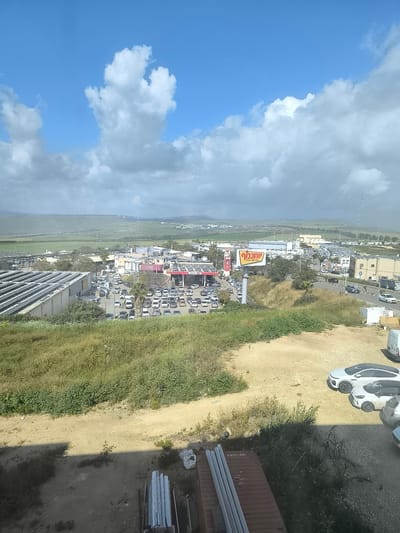 The image depicts a view from an elevated position, possibly a building, overlooking a commercial area in Tiberias, Israel. In the mid-ground is a large parking lot filled with cars, adjacent to what appears to be a shopping complex. A prominent sign with Hebrew lettering is visible within the commercial area. The foreground features vegetation and construction materials (pipes and possibly a container). Several parked cars are also visible in the foreground. The background shows a rural landscape with rolling hills under a sky with scattered clouds. The weather appears overcast. There are no discernible people visible in the image.