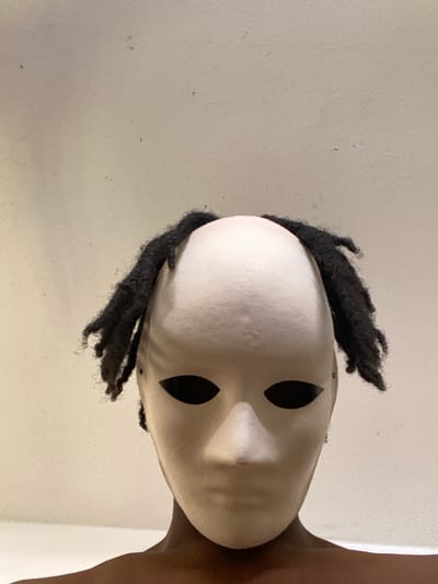 The scene depicts an individual located within an interior space in the city of Capesterre-Belle-Eau, France; however, no specific architectural or environmental markers are present to identify the precise location within the city. The individual is positioned centrally, facing directly towards the viewer. They are wearing a white, smooth, untextured mask covering the entire face from the forehead to the chin, featuring two dark, almond-shaped eye cutouts that reveal no discernible features behind them. Black dreadlocks are visible protruding from around the sides and top of the mask, framing the top portion of the head. Darker skin is visible at the base of the neck and upper shoulders, suggesting the person is bare-chested or wearing dark, form-fitting attire. The background consists of a plain, light beige or off-white wall exhibiting minor surface irregularities and dark specks. Lighting is even, indicating an indoor setting with no distinct light sources or strong shadows.