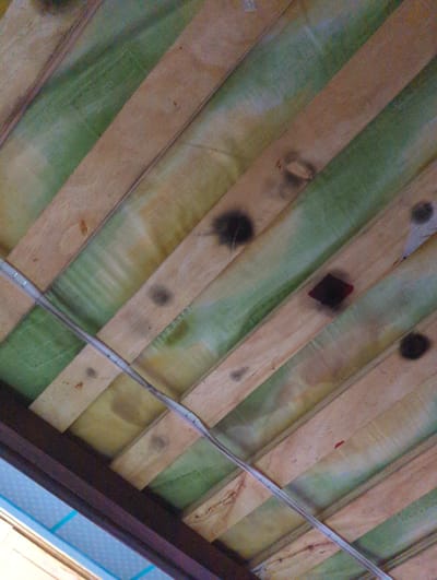The image presents an upward-angled view of a ceiling structure located in the city of Chaguanas, Trinidad and Tobago. The ceiling is composed of parallel light brown wooden planks positioned diagonally across the frame. Between these planks, sections of green and yellowish-green material, potentially insulation or panels, are visible. Numerous irregular dark, circular or oval-shaped stains, consistent with scorch marks or soot, are distributed across both the wooden planks and the green/yellow material. A prominent dark red, diamond-shaped mark, appearing as a localized burn or melted substance, is centered within a larger dark smudge on one of the green panels. A silvery-grey electrical conduit or cable is affixed diagonally across the wooden planks with metal clips or staples. In the lower-left portion of the frame, a dark brown structural beam is present, with a partial view of a light blue patterned surface visible beneath it. No individuals, specific actions, or interactions are depicted.