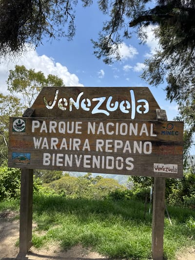 A wooden sign for Parque Nacional Waraira Repano stands near San Antonio de Galipán, Venezuela. The sign, weathered and brown, has the name "Venezuela" cut out in large, flowing letters at the top, with the white painted words "PARQUE NACIONAL WARAIRA REPARO BIENVENIDOS" below. The letters are vertically stacked and off-centered. The sign is partially obscured by trees. A view of the valley below is visible through the cut-out letters. Small logos for "MINEC", "VDD VENTEL", and "INPARQUES" are located near the bottom of the sign. The ground is covered in grass and dirt. The sky is visible through the trees, with blue patches and scattered clouds.