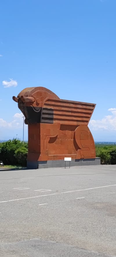 The image shows a large, stylized bull sculpture made of reddish-brown stone. The sculpture is situated outdoors on a paved surface, likely a parking lot or plaza, with white lines marking parking spaces. In the background, there is lush green foliage and a distant landscape with hazy mountains under a bright blue sky with scattered white clouds. The sculpture itself is abstract, with bold geometric shapes forming the bull's head, body, and hindquarters. The head features prominent horns and a dark recess for the face. The body is composed of horizontal bands, creating a textured effect. The sculpture stands on a grey stone base. A small, white rectangular sign is visible in front of the sculpture. The overall scene suggests a monument or landmark located in an open, scenic area. This specific sculpture is known as the "Wind Monument" or "Lion-Bull Monument" and is located in the city of Araks, Armenia.