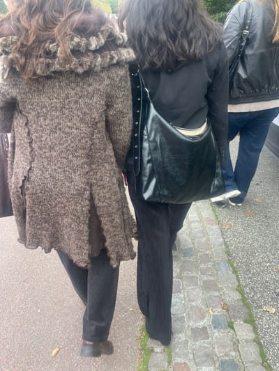 The image displays three individuals walking away from the camera on a street or pathway in Humlebæk, Denmark. The leftmost person, with long, wavy brown hair, wears a brown, textured knit coat with a prominent fur-trimmed collar, dark trousers, and brown low-heeled shoes. The middle person, with long, dark wavy hair, is dressed in a black blazer, dark vertically ribbed trousers, and carries a black glossy leather shoulder bag featuring a studded strap over their left shoulder; a light-colored interior lining is visible. The third individual, partially visible on the far right, wears a black leather bomber-style jacket over a light top, blue denim jeans, and white sneakers. The ground on the left consists of an asphalt surface, transitioning to a cobblestone pavement on the right with visible grass growing between the stones and scattered fallen leaves. The scene is under overcast lighting conditions.