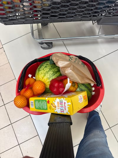 A red shopping basket is filled with groceries, including two watermelons, three oranges in netting, a red pepper in netting, and a yellow box of breakfast cereal. A brown paper bag is also in the basket. The basket is being pushed by a person wearing blue jeans and white sneakers. The scene appears to be in a supermarket in La Trinité, France, indicated by the text on the cereal box, which is in French and mentions "Auchan," a French supermarket chain. The floor is tiled.