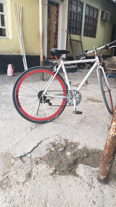A white bicycle with red rims is parked on a concrete surface in front of a building. The bicycle has a black seat, silver handlebars, and silver pedals. The rear wheel has a derailleur. The scene is outdoors, with a yellow wall and a doorway visible behind the bicycle. There are also some white poles leaning against the wall and a pink thermos on the ground. The ground is cracked concrete with some patches of dirt and gravel. A rusty metal pole is visible on the right side of the image.
The time of day appears to be daytime, given the lighting. The weather seems to be fair. The location cues suggest a residential area in Navobod, Tajikistan. There are no people visible in the image, and no specific activity is taking place.
Visible text on the rear tire: "ANT-E".