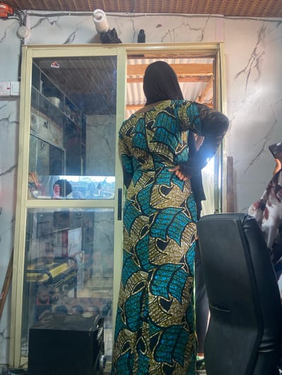 An individual wearing a traditional, patterned dress and a head covering is seen from behind, standing near a glass-fronted display case. The case appears to contain various objects, including packaged goods and what seems to be an electronic device. The surrounding environment consists of a building interior with patterned walls and a view through an open section that reveals wooden beams and framing. The location is Okengwe - Obehira, Nigeria. A black chair with a black cushion is visible. Electrical wiring and fixtures are visible. An Adidas sticker is placed on the left side of the glass-fronted display case.