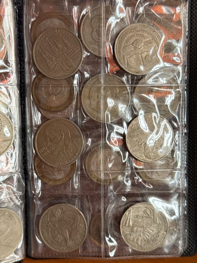 The image displays a page from a coin album. The page is divided into clear plastic sleeves, each containing one or more coins. The coins are mostly round and metallic, with various engravings and inscriptions. Many of the coins appear to be from the Soviet era, featuring prominent figures like Lenin and symbols such as the hammer and sickle. Some coins are dated, including 1980, 1970, and 1981. The album itself is black and appears to be bound. The background is out of focus but suggests an indoor setting, possibly a room. There are no people or discernible actions taking place. The overall impression is one of a collection of historical currency. The image is a close-up shot, focusing on the contents of the coin album.