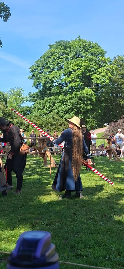 In Turku, Finland, on a sunny day, a reenactment event is taking place. Two individuals, dressed in historical attire, are engaged in a jousting practice session with wooden poles. The person on the left wears a black tunic and hat, while the person on the right is adorned in a blue dress and a straw hat. They are surrounded by an audience of onlookers, some seated and others standing, observing the activity. The event is set in a grassy field with lush green trees in the background, under a clear blue sky.