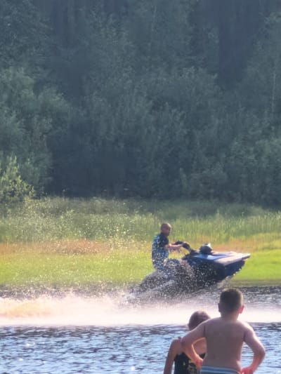 In the foreground of the image, two young children are standing in the water, with their backs to the camera. The child on the right is wearing striped swim trunks, and the child on the left is wearing a dark swimsuit. In the middle ground, a person is riding a jet ski, creating a large spray of water as they move. The jet ski is dark blue. The background consists of a dense forest of trees, with a field of green grass and scattered wildflowers in front of it. The scene appears to be a recreational area, likely a lake or river, given the presence of water and the activities depicted. The image does not contain any specific landmarks that would allow for pinpointing the exact location within Sysert, Russia.