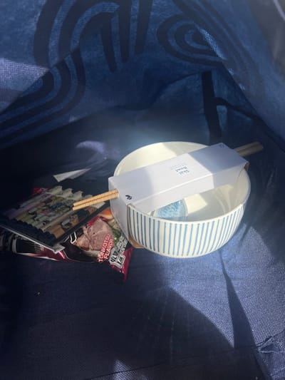 The image displays the interior of a dark blue fabric bag, set within the Hof Ashkelon Regional Council, Israel. The bag's inner lining features a repeating pattern of darker blue curved lines. Strong, direct light from the upper right illuminates the contents, casting shadows to the left.
Within the bag, two ceramic bowls are stacked centrally. The upper bowl is plain white. The lower bowl is white with vertical light blue stripes. A white cardboard sleeve labeled "Skal Bowl" is positioned horizontally across the top of the stacked bowls, securing a pair of light-colored wooden chopsticks. A small, light blue spherical object is visible inside the bottom bowl.
To the left of the bowls, a red and white packet marked "Tonkotsu Flavor" (indicating instant ramen) is partially visible. Below this packet is a stack of several flat, rectangular items displaying decorative patterns. One of the chopsticks rests across the top of this stack of items. No people are present.