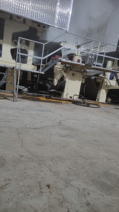 The image depicts a complex industrial machinery setup. Metal railings and structures are prominently featured, partially obscuring the core machinery. A man, seemingly working on the equipment, is visible. The machinery is positioned on a concrete floor, which shows signs of cracking and wear. A wheel or circular object lies near the machine, with a yellow stripe painted near the equipment on the floor. The lighting appears to be a combination of natural and artificial sources.
