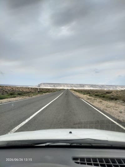 This is a first-person perspective, point-of-view shot from a vehicle driving on a paved road in a desert landscape. The road has two lanes, marked with white lines. The terrain on either side of the road is arid, with sparse, dry vegetation and patches of sand. In the distance, a long, undulating ridge of white, rocky cliffs dominates the horizon. The sky is overcast with heavy, grey clouds, suggesting a potentially gloomy or stormy day. The time is displayed as 13:15 on June 26, 2026. There is a single car visible in the distance on the road. The overall mood is one of travel through a desolate and remote environment. The location is identified as Saiyn Şapağatov, Kazakhstan.