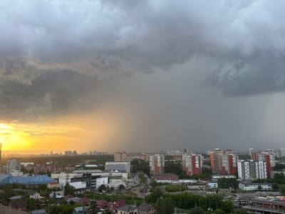 The image shows an aerial view of a city skyline with a dramatic sky. The left side of the sky is illuminated by a bright, golden sunset, casting a warm glow over the landscape. On the right, the sky is dominated by dark, heavy storm clouds, creating a stark contrast.

In the foreground, a dense urban area is visible, featuring a mix of residential buildings, apartment complexes, and commercial structures. The apartment buildings are predominantly tall and modern, with some exhibiting colorful facades. Various trees and green spaces are interspersed throughout the city.

The overall scene suggests a transition from a clear, sunny day to an impending storm. The location is identified as Shirkovo, Russia. There are no visible people in the image.