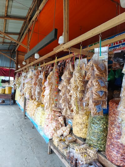 The image depicts a stall selling a wide variety of snacks, likely in Sawang, Indonesia. The stall is set up outdoors, under a wooden structure with an orange tarp for a roof, suggesting a market or roadside vendor. Various snacks are displayed, mostly in clear plastic bags, hanging from hooks or arranged on shelves and tables. These include an assortment of chips, crackers, nuts, and what appear to be puffed rice snacks, with colors ranging from pale yellow and white to green and reddish-brown.
Some of the snacks are packaged in large, clear plastic sacks, while others are in smaller, individually sealed bags. Several bags have labels with text, though most are too small or blurry to be read clearly. One prominent label, visible on several bags of triangular-shaped snacks, features a blue design with white text. Large bags of light yellow, green, and reddish-brown snacks are particularly noticeable due to their size.
In the background, there are glimpses of corrugated metal siding and some greenery, hinting at the surrounding environment. A long, black digital display board is mounted above the stall. The lighting suggests it is daytime, possibly late morning or afternoon, with diffused sunlight. The overall impression is that of a bustling market scene with a vibrant display of local food products. There are no people visible in the immediate foreground or actively engaged in transactions, focusing the view solely on the merchandise.