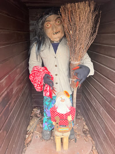 A Halloween decoration depicting a witch is positioned inside a wooden structure, possibly a shed or storage box, in Sanatoriya Yumatovo Imeni 15-Letiya Bassr, Russia. The witch figure has a grotesque mask with yellowed teeth and bulging eyes, black and grey hair, black gloves with long, red, glittery nails, and a beige jacket over a blue shirt. A red and white polka dot kerchief is also visible. The witch holds a brown twig broom in one hand. To the right of the witch is another doll or figure with a red and white polka dot outfit and a sunflower accent. The doll sits on a small wooden structure with some Russian writing visible. Pine cones and leaves surround the base of the doll. The wooden structure has a brown, slatted interior.