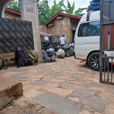 The image depicts an outdoor staging area in Mweka, Tanzania. In the foreground, the ground is paved with irregular flagstones in varying shades of brown and grey. A rough concrete block lies in the lower-left corner. To the left, a thick concrete pillar with an arched opening stands, against which a large black sheet with "ZURKT" branding is propped.

In the midground, a significant quantity of luggage, consisting of numerous large backpacks and duffel bags in colors such as green, black, and blue, is piled on the ground. A large, light-colored, irregularly shaped object, possibly a rock or wrapped bundle, is visible among the bags. Two three-wheeled auto-rickshaws (Tuk-Tuks) with black roofs are parked side-by-side behind the luggage, also appearing to carry items.

Several individuals are present. One man wearing a white t-shirt and blue cap stands near the auto-rickshaws, facing away from the viewer. Another man in a white t-shirt is seated nearby. A white van, possibly a passenger model, is parked on the right, partially visible. Its roof rack holds blue objects, and at least one person is visible through its tinted windows. An individual is seated near the van's rear. A partially open metal gate occupies the far right side of the frame.

The background features stone-clad buildings with reddish-brown roofs. Lush green banana trees and other tropical foliage are visible behind these structures under an overcast sky. The overall scene suggests a transit point or a preparation area for travel, characterized by packed luggage and parked vehicles.
