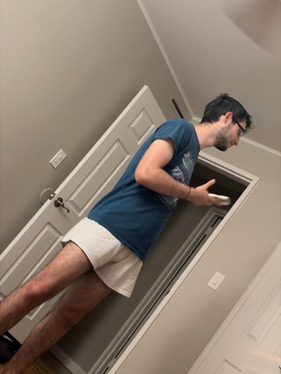 A young man is captured in an indoor setting, likely a hallway or room in a residential building in Franklin Township, United States of America. He has dark hair, wears glasses, and has a beard. He is dressed in a blue short-sleeved t-shirt with a graphic design and light-colored shorts, exposing his legs.
The man stands, holding a light-colored smartphone in his right hand, and is looking upwards and slightly to his right, his expression neutral. The background features a light grey or beige wall with white ceiling molding and a white paneled door. The door has a dark-colored handle with a ring hanging from it. A white light switch is visible on the wall to the left of the door. The image is taken from a low angle, giving a tilted perspective of the scene. The lighting appears to be typical indoor ambient light, and no specific time of day is discernible from external cues. There is no visible text in the image.