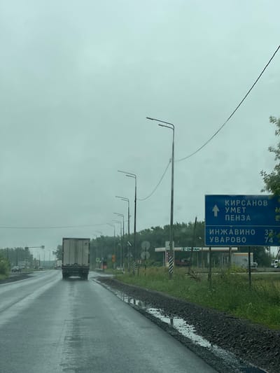 An overcast, possibly rainy day. A paved road curves slightly to the left, with a wet surface reflecting the sky. A large, white-sided truck with a dark tarp covering its load is in the right lane, traveling away from the camera. To the right of the road, there is a grassy area with a few trees and a blue road sign. The sign indicates directions to "КИРСАНОВ УМЕТ ПЕНЗА" with an upward arrow and "ИНЖАВИНО 37" and "УВАРОВО 74" below. Further to the right, a gas station with "OKTAN" branding is visible, along with other vehicles. Several lampposts are positioned along the right side of the road. The environment appears to be rural or on the outskirts of a town. There are no visible people.