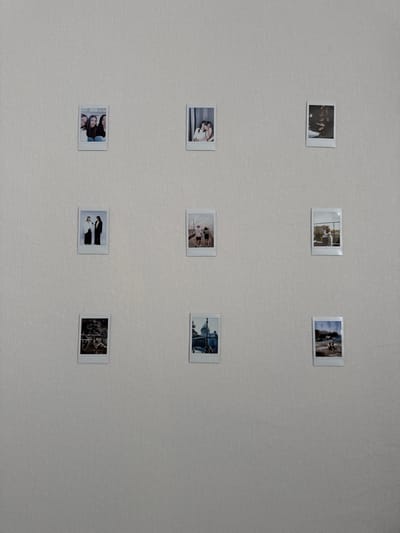 Nine small, square photographs are arranged in a 3x3 grid on a light, textured wall. The photographs appear to be Polaroids or similar instant prints. The content of the photographs is varied and includes images of people, possibly in groups or couples, as well as images of architecture or scenery. The wall is a neutral color and appears to be made of a textured material like wallpaper. The overall lighting of the scene is diffused and even. The photos do not appear to be particularly new. Location unspecified.