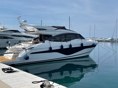 A medium shot captures a docked luxury yacht in Grad Rovinj, Croatia. The yacht, predominantly white with black accents, is moored at a concrete pier. Dark blue fenders are strategically placed along its hull. A section of teak decking is visible at the stern. Mooring lines secure the yacht to metal bollards on the pier. The water reflects the yacht and surrounding environment, creating a mirrored effect. In the background, other yachts are docked, and their masts punctuate the skyline. The sky is light blue with scattered clouds.