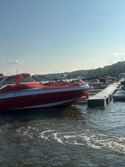 A fleet of boats is moored at a dock on a body of water under a clear sky with scattered clouds. The boats, mostly covered with red and black tarps, are lined up along a wooden pier. One red and white speedboat is prominently featured in the foreground. The water exhibits a slight ripple effect, with some areas of foamy churn. Lush green trees and hills form the background, suggesting a natural setting. The scene is likely in Algonquin, Illinois, a city known for its picturesque waterfront and recreational opportunities.