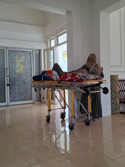 A person in striped clothing is lying on a foldable medical stretcher, viewed from a high angle. The stretcher is indoors on a tiled floor in a building with a large window. The location is Uleereuleung, Indonesia.