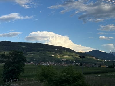 The image depicts a landscape view with a village nestled in a valley, surrounded by lush green hills and mountains under a partly cloudy sky. The village, identified as Nave San Rocco, Italy, features buildings with terracotta roofs. The hills are covered with vegetation and vineyards, indicating agricultural activity. In the foreground, there are fields and trees. The lighting suggests it is either early morning or late afternoon, with sunlight casting shadows and highlighting certain areas of the landscape. The overall scene conveys a sense of rural tranquility.