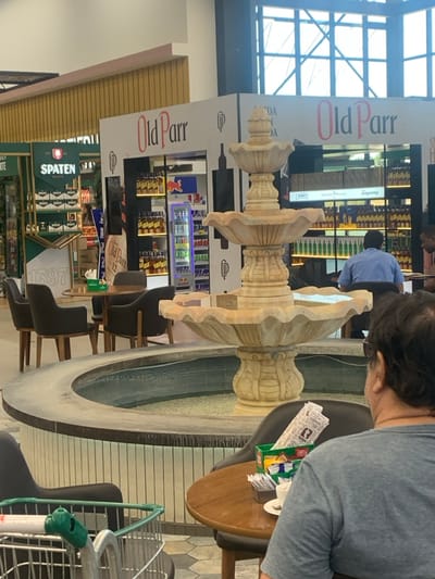 This image depicts the interior of a large retail establishment, specifically a hypermarket, located in Lauro de Freitas, Brazil.
The central feature is a three-tiered, light-colored ornate stone fountain with water contained in its basin, surrounded by a low, vertically-striped tiled wall. In the immediate foreground, a male individual is seated with his back to the viewer, wearing a grey t-shirt and sunglasses, at a round wooden table. A green container with wrapped utensils and a white cup are on his table. A silver shopping cart with green handles is partially visible to the left.
In the midground, a seating area with multiple dark-upholstered chairs and round wooden tables is present. Beyond this, two prominent display kiosks branded "Old Parr" are visible, showcasing multiple shelves stocked with amber-colored liquor bottles. One "Old Parr" kiosk includes a vertical refrigerated display unit containing various canned beverages, notably a Red Bull can. To the far left, a green "Spaten" branded display featuring shelves of bottles is partially obscured, alongside a sign displaying the number "1397." Another individual, dressed in a blue shirt, is seated at a table within the liquor section in the background to the right.
The environment features high ceilings, large windows allowing natural light in the upper background, and a tiled floor with a hexagonal pattern. The architecture suggests a modern, expansive retail space. Based on the prominent brand displays and architectural characteristics, this scene is situated within a hypermarket, likely a G Barbosa Hiper store, in Lauro de Freitas, Brazil.