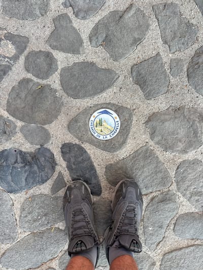 The image shows a close-up view of a cobblestone path in Moustiers-Sainte-Marie, France. A circular sign with an illustration of a village and mountains is embedded in the ground. The sign reads "CHEMIN DE LA CHAPELLE" and "MOUSTIERS S-STE-MARIE". A pair of grey athletic shoes and a portion of a person's legs are visible at the bottom of the frame, indicating the viewer is standing on the path. The stones are irregular in shape and size, and are set in a grey concrete mortar. There are also small pebbles and a few scattered leaves on the ground.