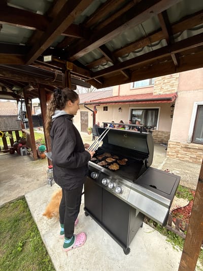 A woman in a black jacket and pants, wearing pink and green Crocs, is grilling meat on a large stainless steel grill outdoors in Vinarovo, Bulgaria. She uses tongs to flip the meat. A golden dog is lying near her feet. The grill is on a concrete patio with grass and foliage surrounding it. In the background, people are sitting at a table on a covered porch. A wooden pergola covers the immediate foreground. The grill is situated in front of a house.