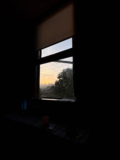 The image depicts a window looking out onto a misty sunrise over a landscape of trees and buildings, possibly in Jāņupe, Latvia. The room is dark, with light streaming in from the window, casting shadows on the floor and a surface below the window. On this surface, a blue cylindrical object, an orange cup, and a pack of cigarettes are visible. The window frame is dark wood, and a light-colored blind is pulled down at the top of the window. The sky is filled with soft, warm light, and the mist obscures the details of the landscape, creating an atmospheric and serene scene.