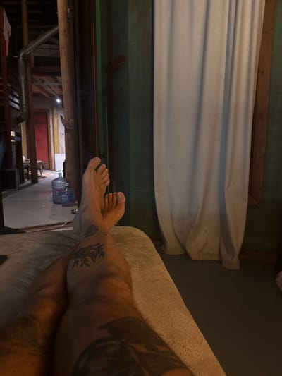 A person reclines on a light beige surface. Their legs are visible, adorned with tattoos. The background reveals a scene of exposed wooden beams and a glimpse into an adjacent room featuring a red door. Curtains hang above, contrasting with the darker walls and woodwork. Water bottles are placed along a wall.