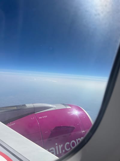 The image is a photograph taken from an airplane window, showing a view of the earth below and part of the airplane's engine. The scene is dominated by the vast expanse of the sky, transitioning from a deep blue near the top of the frame to a lighter, hazy blue closer to the horizon. A layer of clouds is visible on the horizon, creating a distinct line between the sky and the earth. The earth's surface appears flat and featureless from this altitude, with a faint, hazy texture.
The most prominent object in the frame is the airplane's engine, painted in a vibrant magenta color. The engine nacelle is detailed, with visible panels and warning labels. Text is visible on the engine, including "NO STEP," and "wizzair.com" in large white letters on the magenta portion. The sun glints off the surface of the engine, creating a bright highlight. The airplane's wing, painted white with red and white stripes, is visible in the lower right corner of the frame, obscuring a small part of the view.
The image appears to be taken during the daytime, with bright sunlight casting shadows and highlights. The weather seems clear, with the sun shining brightly, though there is some haze at lower altitudes. The perspective suggests the airplane is in flight at a high altitude, likely cruising. There are no people visible in the image. The overall mood is one of travel and the vastness of the sky. The location context indicates the flight might be over or near Obrenovac Urban Municipality, Serbia, though this cannot be definitively confirmed from the visual information alone.