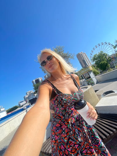 A woman with blonde hair and sunglasses is taking a selfie outdoors, holding a white cup with a straw. She is wearing a colorful, patterned dress and standing on a paved area next to a wooden bench. In the background, there are several tall buildings, a Ferris wheel, trees, and a blue sky. The location appears to be a city waterfront. The text on a kiosk in the background is partially visible and includes the word "ЭКСКУРСИЯ," which means "excursion" in Russian. This suggests the location might be a tourist area.