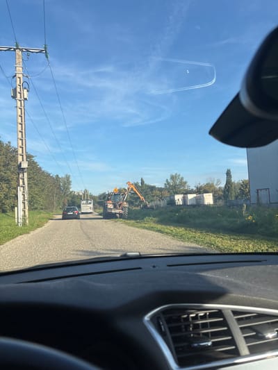 The image captures a daylight scene from within a vehicle in Scherwiller, France, looking forward down a narrow, paved road. The sky is predominantly blue with sparse white clouds and two distinct airplane contrails in the upper right, one forming a U-shape, within which two small circular reflections or objects are visible. A black object, likely a vehicle interior component, partially obstructs the upper right sky.

In the foreground, the dark dashboard and a segment of the steering wheel are visible, along with air vents on the right. A utility pole with power lines and a transformer stands vertically on the left side of the road. To the left of the road, dense green foliage and trees line the path, with a multi-story building faintly visible through them in the distance.

On the right side of the road, an orange and black tractor-like vehicle, equipped with an extended articulated arm featuring a cutting or trimming attachment, is positioned on the grassy shoulder, facing away from the camera. A yellow triangular warning sign with a black exclamation mark is affixed to its rear. Further down the road, a black sedan is stationary, followed by a white bus. The immediate area to the right of the road features green grass and vegetation. Beyond the tractor, several white, container-like structures are visible in a semi-industrial setting, adjacent to a large, light-colored building with a dark roof. Scattered trees, including slender cypresses, populate the background. No individuals are explicitly visible, but the presence of vehicles implies occupants.