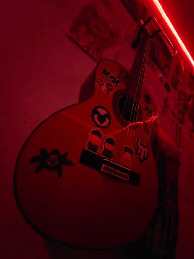 The image displays an acoustic guitar resting in a setting illuminated almost entirely by a vivid red light. The entire scene, including the guitar and its surroundings, is saturated with deep red and dark red tones.
The main subject is a light-colored acoustic guitar, which appears bright red due to the pervasive lighting. Its body is adorned with several stickers: a prominent "AC/DC" logo (white letters on a black background, now appearing red and dark against the red guitar body), a circular black sticker containing a stylized red symbol (resembling a peace sign or an anarchy symbol), and a dark, possibly black, sticker resembling a starburst or floral design on the lower bout. Another smaller, indistinct sticker is visible near the "AC/DC" logo, and a stylized letter or symbol is on the upper bout closer to the sound hole. The guitar's sound hole, dark bridge, and strings are clearly visible, stretching towards the fretboard and the partially visible headstock with its tuning pegs.
The dominant light source is a bright strip of red LED lights running along the top-left edge of the frame, casting the intense crimson hue over the entire image. The guitar is resting on a flat surface, which appears light in tone but is rendered red by the lighting. In the background, behind the guitar and to its left, there are indistinct items that appear to be papers, posters, or flat objects, also bathed in the red light. One faint image, possibly a stylized portrait or drawing, is barely discernible on a background item to the left of the guitar's neck.
No specific activity is taking place; the guitar is stationary. The strong red lighting creates a dramatic and possibly moody or energetic ambiance, suggesting an indoor setting. The only clearly visible text is "AC/DC" on one of the guitar stickers.