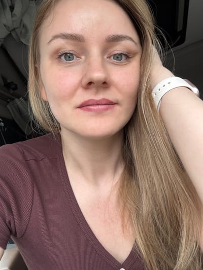 A close-up, selfie-style shot of a fair-skinned woman with blonde hair. Her right arm is raised near her head, a white smartwatch visible on her wrist. She is wearing a maroon-colored, crew-neck top. Her face is visible, with prominent eyebrows, grey eyes, and lips painted in a mauve shade. The background is out of focus, appearing to be indoors, potentially near a car. There are no indications this photo was taken in Kukui, Russia.