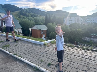 A man and a boy stand on a paved pathway in a mountainous area. The man, wearing a light-colored t-shirt with text and dark shorts, is positioned on the left, looking down and pointing. The boy, in a light-colored t-shirt and dark shorts, faces forward and extends his right hand. Behind them, buildings with green roofs and a wooden structure are visible, nestled amongst lush green trees. The background features rolling hills and a bright, partly cloudy sky. The location is identified as Danilovka (Altai), Russia.