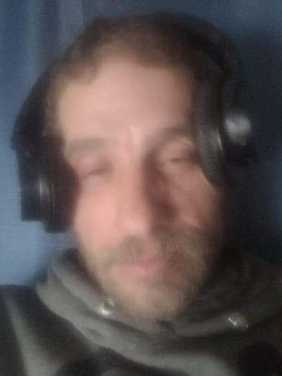 The image presents a blurred, head-and-shoulders portrait of an adult male. The subject has dark hair, visible thinning on the scalp, and a brown beard covering the chin and jawline. He is wearing black over-ear headphones and a grey hooded sweatshirt, with a single silver-toned grommet visible on the garment near the neckline. The subject's eyes are closed. The background is a uniform, indistinct blue, indicative of an indoor setting. No specific visual cues are present to determine a precise location within Medicine Hat, Canada.