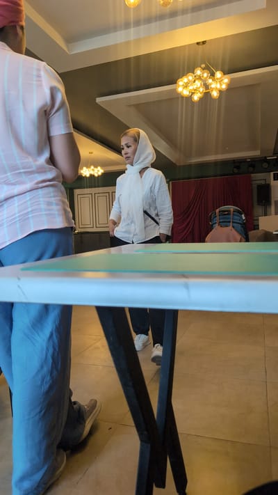 In a room with cream-colored walls and a dark green ceiling, two individuals are present. The person in the foreground, on the left, is seen from the back, wearing a pink and white striped short-sleeved shirt and blue pants. Their head is covered with a pink turban. They are standing near a white table with a green surface, positioned in front of a black metal-legged table.

To the right, a woman is facing left, looking downwards. She is wearing a white headscarf and a white long-sleeved shirt, tucked into dark pants. She has a black purse slung across her shoulder. Her hands are in her pockets. She is standing on a tiled floor. In the background, red curtains hang, and a bag with blue denim and a tan leather strap is placed on a chair. The room is illuminated by a chandelier with multiple light bulbs. The location is identified as Ötegen Batyr, Kazakhstan.