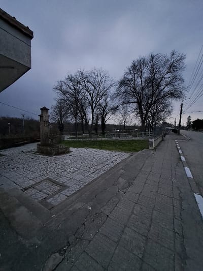 The image depicts an outdoor scene in Kamenets, Bulgaria, under an overcast sky with dim, diffused lighting. In the foreground, the lower left corner shows the underside of a concrete architectural overhang. To its right, a dark grey paved area, composed of irregularly shaped stone pavers, occupies the mid-ground. Centrally positioned within this paved area is a multi-tiered stone monument, likely a commemorative cross or pillar, featuring an inscription on its front face. Beyond the paved area, a patch of green grass is visible, bordered by a low concrete curb. Behind the grass, a line of mature, bare deciduous trees stands prominently, their dark branches silhouetted against the sky. Further bare trees are visible in the distant background. To the right of the paved area, a damaged, dark grey sidewalk with a distinct pattern of cracks runs parallel to a paved road, which is marked by a white line. A utility pole with multiple overhead wires is situated on the far right, adjacent to the road. No individuals are present in the scene, nor are any actions or interactions depicted.
