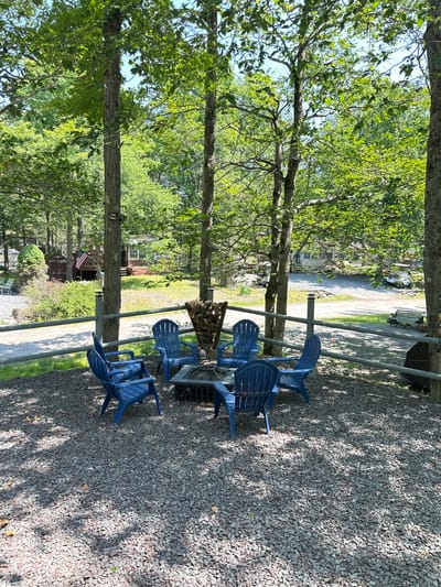 In Covington Township, United States of America, an outdoor fire pit area is depicted. Seven blue Adirondack chairs are arranged in a circle around a square metal fire pit filled with logs. The ground is covered with dark gray gravel, and dappled sunlight filters through the overhead trees. A grey wooden fence encloses the area. In the background, there are several mobile homes and a building with an American flag flying, suggesting a residential or recreational park setting. The trees are lush green, indicating a warm weather season.