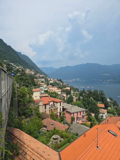 The image captures a scenic view of a hillside village nestled beside a lake, with mountains in the background. The perspective is from a high vantage point, looking down at a densely populated area characterized by terracotta-roofed buildings and lush greenery. The village appears to be built on a steep incline, leading down towards a body of water, likely a lake, with a few boats visible on its surface.

Prominently in the foreground are several buildings with orange, tiled roofs, some featuring skylights and chimneys. The architecture suggests a Mediterranean or European style, with pastel-colored facades and balconies. Dense foliage, including trees and shrubs, surrounds the houses, indicating a warm climate and well-maintained gardens.

Further into the scene, the village sprawls across the hillside, showcasing a variety of architectural styles and colors. A red and white construction crane is visible in the distance, near the lake's edge, suggesting ongoing development or repair work. The majestic, blue-tinged mountains rise in the background, partially obscured by haze, contributing to the depth of the landscape.

The sky is a clear, light blue, with a few scattered white clouds, indicating fair weather and likely daytime. A blue sign with white symbols, possibly a directional or informational marker, is visible on a metal fence in the mid-ground, suggesting a public pathway or viewing area. There is no indication of people or specific activities, lending the scene a peaceful and picturesque quality. The overall impression is one of a serene, sun-drenched Italian village overlooking a beautiful natural landscape.