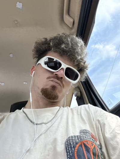The image depicts a close-up, selfie-style shot of a person inside a vehicle. The individual is wearing white, wrap-around sunglasses that obscure their eyes, a light-colored t-shirt with a decorative pocket on the left chest featuring orange and blue stitching, and a silver chain necklace. White wired earbuds are visible, with one earbud in their ear and the wire extending down. The person has curly, light-brown hair and a small, pointed goatee. Their expression is neutral to slightly stern. The interior of the vehicle is visible, including the headliner and a portion of the car door, suggesting a standard passenger car. The sky and clouds are visible through the window. The image does not provide any specific indicators of its location within Bang Rak Noi Subdistrict, Thailand, as the environment depicted is generic to an indoor vehicle setting.