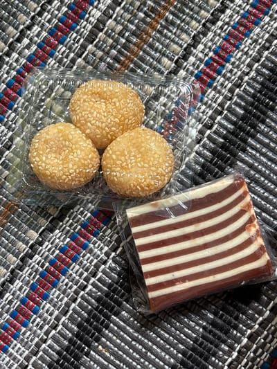 Three sesame seed balls rest in a clear plastic container, positioned adjacent to a rectangular, wrapped piece of striped chocolate and white confection. They sit atop a woven mat featuring a crisscross pattern in black, white, blue, and red hues. The packaging is transparent.