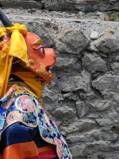 The image shows a close-up of an individual wearing a vibrant, ornate costume and an elaborate mask. The mask is orange with gold detailing around the eyes and mouth, and appears to depict a mythical creature, possibly a dragon or a lion, common in traditional performances. A yellow cloth is draped around the mask, and the costume features rich blue fabric with intricate patterns in red, green, and gold. The background is a rough, gray stone wall. The focus is on the detail of the costume and mask, suggesting a cultural or religious performance is taking place. The lighting suggests daytime, but the weather is not discernible. The setting appears to be outdoors, given the stone wall which could be part of a building or a natural rock formation. The image is cropped to emphasize the performer, with the stone wall occupying the right half of the frame. There is no visible text in the image.