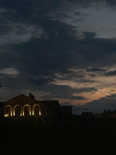 The image depicts the exterior of a building with arched windows illuminated by warm light, situated in the village of Sasunik, Armenia. The sky above is dark and cloudy, with hints of orange and pink suggesting the time is either dawn or dusk. The lower portion of the image is largely dark, obscuring the ground and surrounding environment. The building itself appears to be a house or a similar structure with a sloped roof and chimneys. Some other structures are faintly visible in the background. The overall scene conveys a sense of quietude and a touch of dramatic atmosphere due to the cloudy sky.