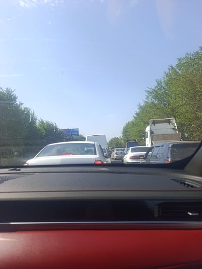 The image shows a traffic jam on a sunny day, likely in or near Ötegen Batyr, Kazakhstan, as indicated by the presence of trees lining the road and a billboard with Cyrillic script. The perspective is from inside a vehicle, looking forward through the windshield. The dashboard of the car is visible in the foreground, with a red element at the bottom.

Multiple vehicles are stopped in traffic, including passenger cars and at least one white truck. The vehicles are positioned on a paved road, with a lane divider visible. Trees form a green barrier along the right side of the road, and more trees are visible on the left. The sky is blue with some scattered white clouds. A billboard is visible in the distance on the left side of the road, featuring text in Cyrillic. The overall scene suggests a typical urban or suburban traffic congestion on a clear day.