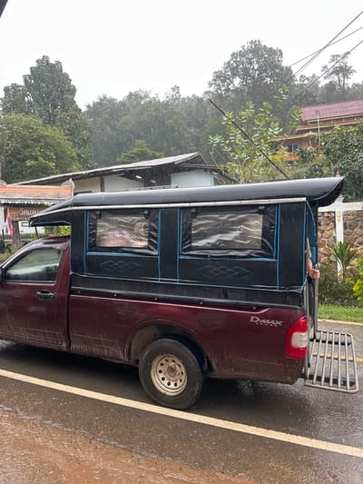 The scene depicts a maroon Isuzu D-MAX pickup truck parked on a wet asphalt road in Ban Paem Bok, Thailand, under a steady rainfall. The truck's bed is equipped with a custom black canopy featuring three clear plastic windows on its visible side, outlined with blue trim. The lower body of the truck appears splattered with mud or dirt. A metal, grid-like ramp or step is lowered from the rear tailgate of the vehicle. A person's arm and a portion of their upper torso, clad in a light-colored shirt, are visible holding onto the internal frame of the canopy on the passenger side. The road surface is visibly wet with reflections, and a solid white line runs parallel to the truck. In the background, dense green foliage covers hillsides under an overcast sky. Several buildings are visible, including one with an ornate, multi-tiered orange roof (possibly a temple) nestled among the trees on an elevated terrain, and another with a simpler, light-colored roof lower down. Utility lines cross the upper portion of the frame. A stone wall with decorative elements is partially visible to the right of the truck. A sign with Thai script and a Thai flag are partially visible to the left.