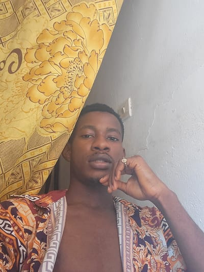 This image depicts an adult male positioned centrally in an indoor setting within the city of São Félix do Xingu, Brazil. The male subject, with dark skin, short dark hair, and light facial hair, is looking directly at the viewer. He wears an unbuttoned, short-sleeved collared shirt over his bare torso, featuring a complex pattern of orange, white, purple, and brown scrollwork and geometric shapes. His left hand is raised to his chin, with a gold-colored ring visible on his left ring finger. The background on the left consists of a yellow fabric curtain displaying large floral and geometric patterns. The background on the right is a light gray wall with visible surface imperfections, including faint cracks and peeling paint. A white electrical outlet or switch plate is affixed to the upper right section of this wall.